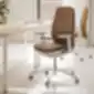 Brūns ergonomisks biroja krēsls ar baltu pamatni un riteņiem blakus gaišam koka galdam modernā telpā, kas piepildīta ar dabisko gaismu.